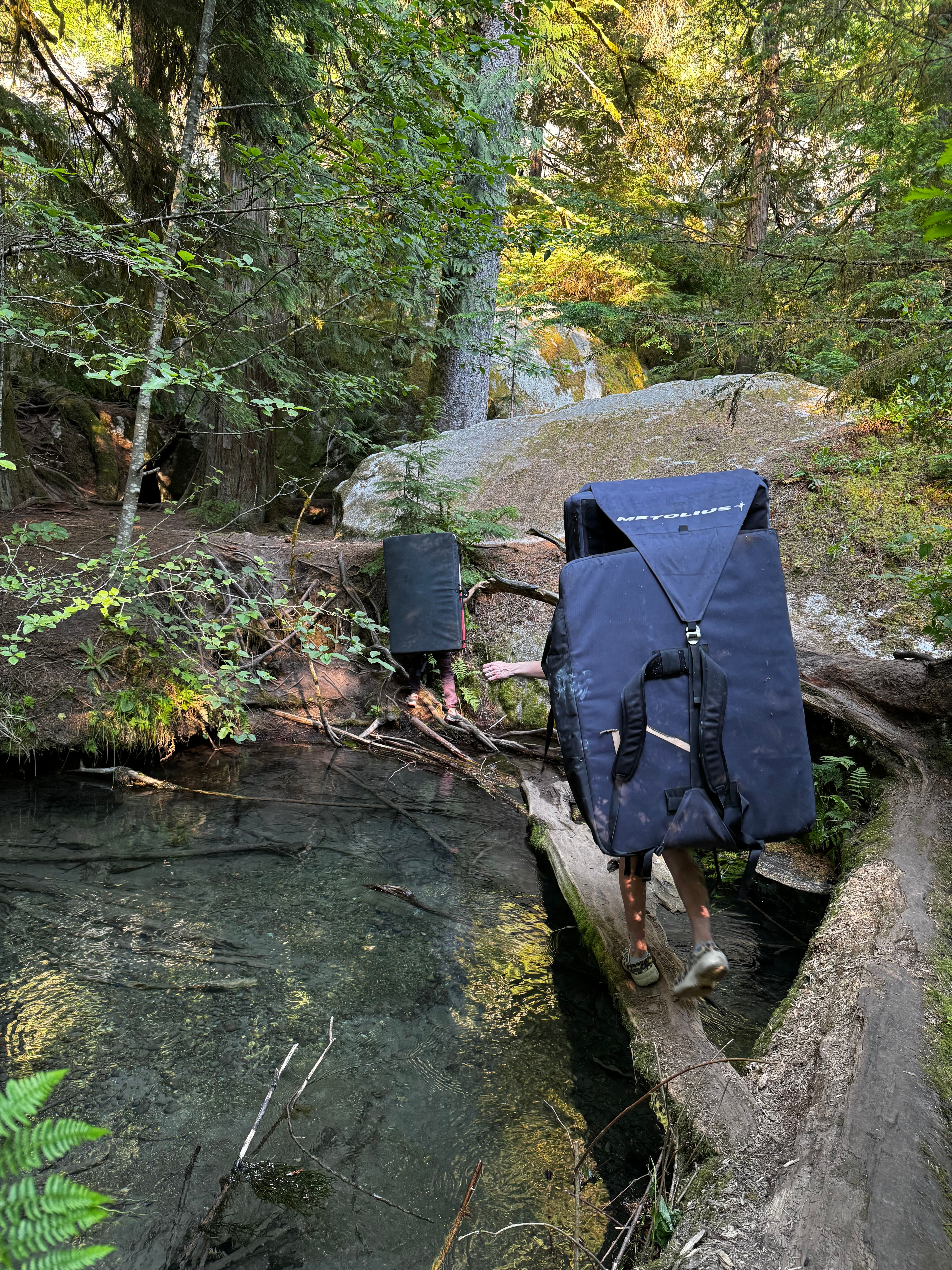 Two people crossing a log over a creek carrying crash pads
